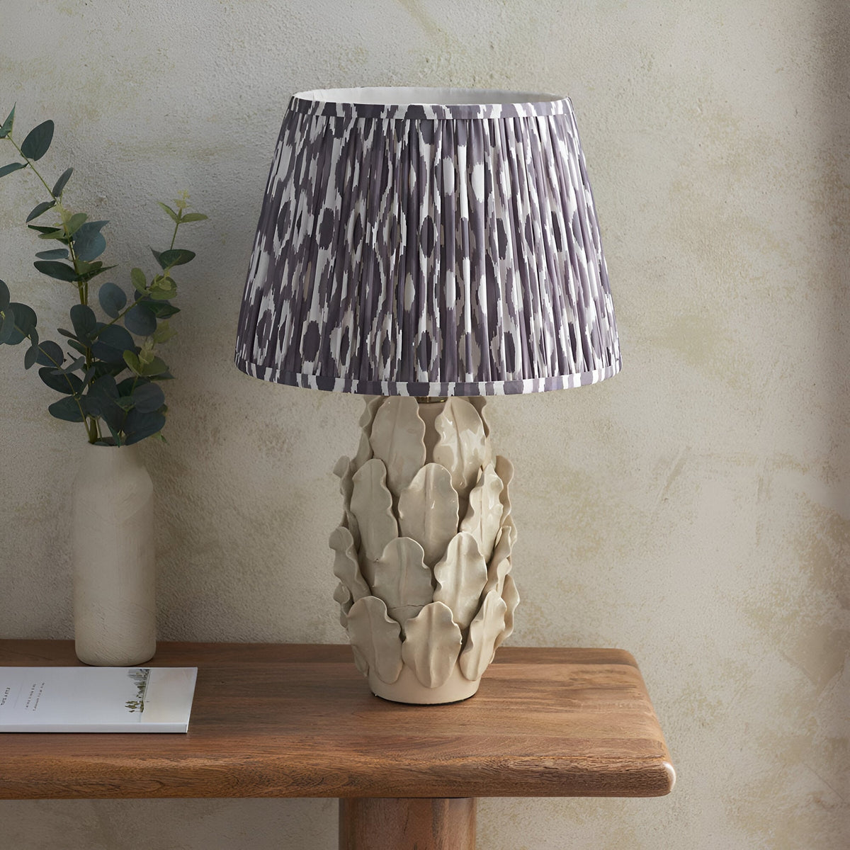 Endon Caldford Ikat 35cm table lamp with ivory crackle-glaze ceramic leaf base and grey pleated Ikat shade, on a wooden console with eucalyptus.