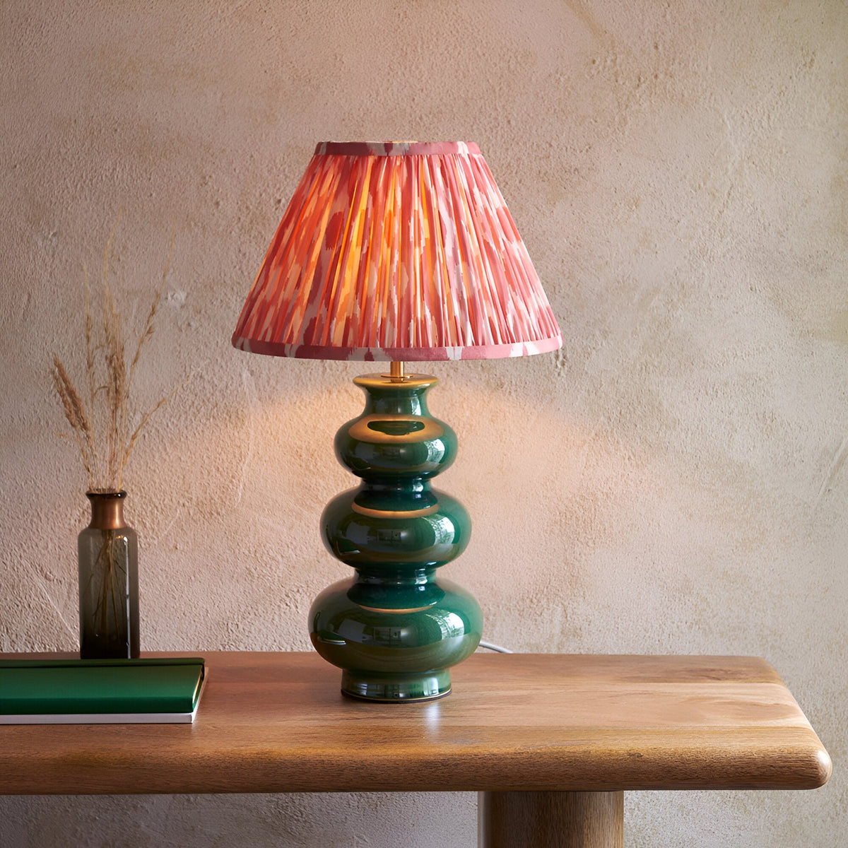 Carlstead Ikat Table Lamp in dark green glossy ceramic with pleated pink and orange patterned shade, shown on a wooden console table.