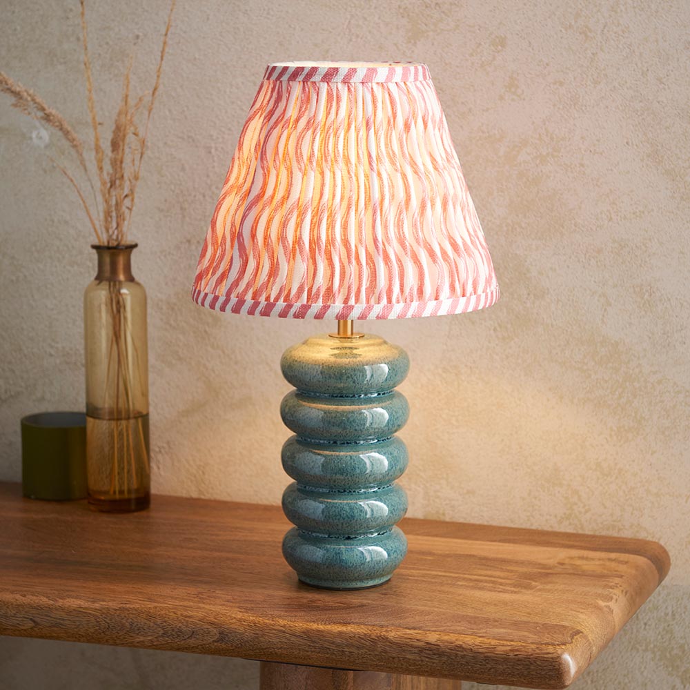 Cliffstead Table Lamp with ribbed teal ceramic base and pink wave-patterned fabric shade, brass fitting, shown lit on a wooden table.