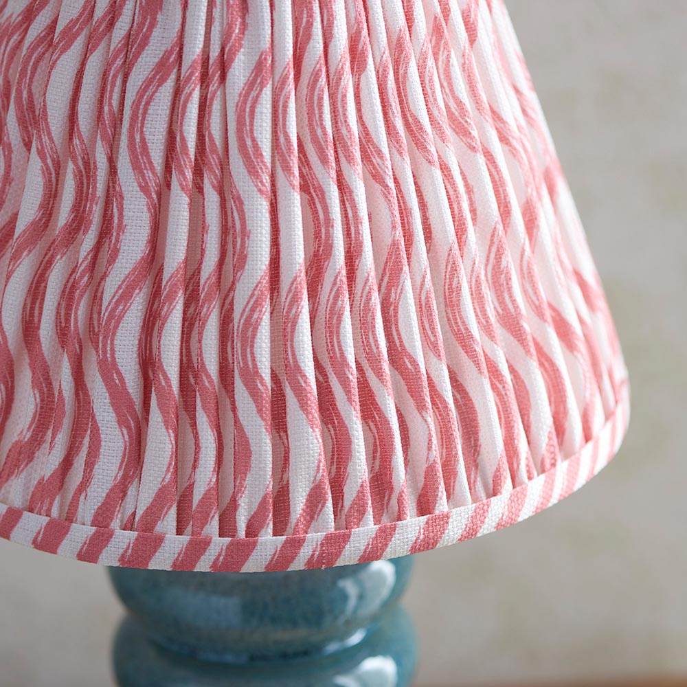 Cliffstead Table Lamp close-up showing pleated pink and white wave-print fabric shade with taped edge, atop a curved ceramic base in a blue reactive glaze.