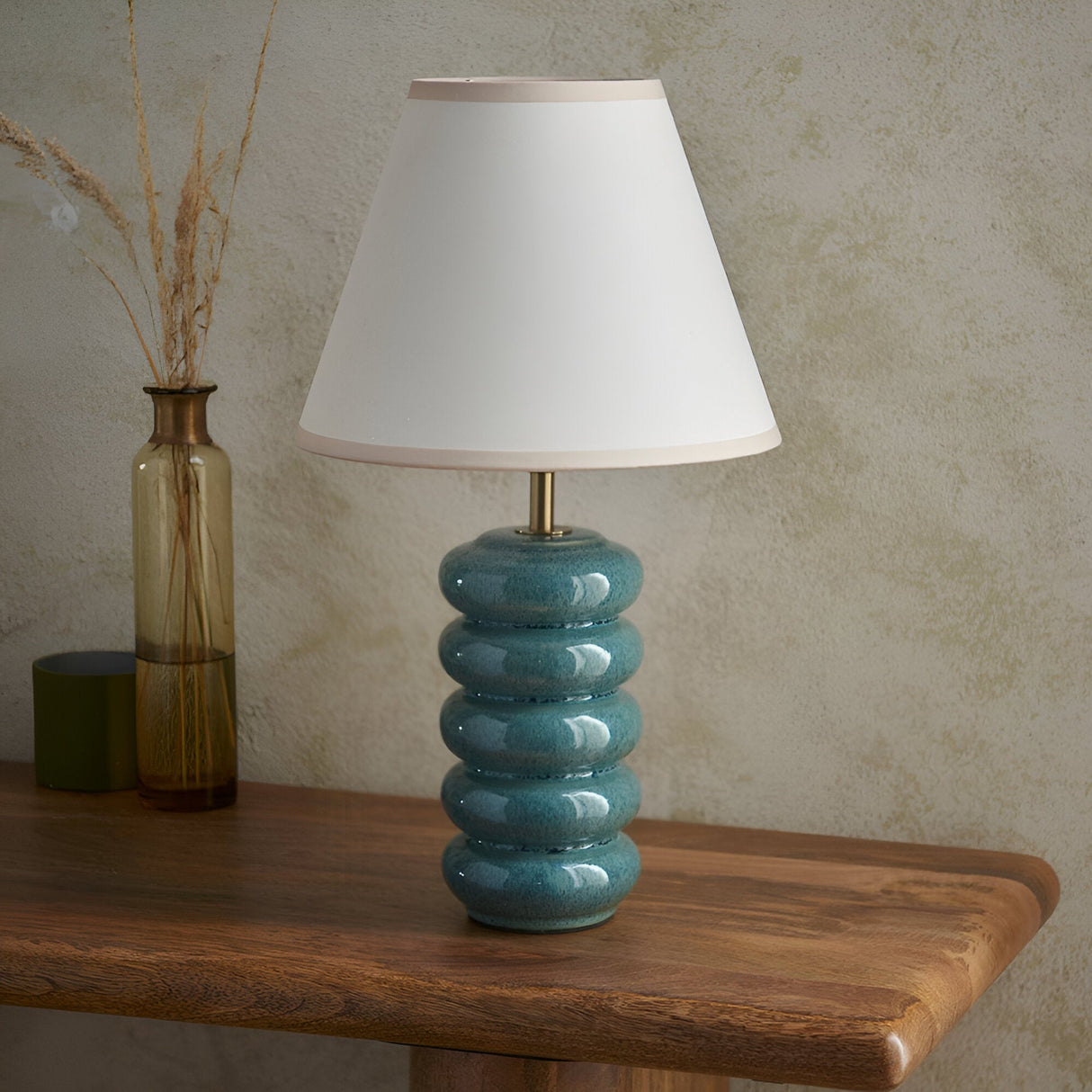 Endon Cliffstead table lamp with stacked ocean spray blue-green ceramic base, antique brass stem and ivory tapered shade, on a wood console beside a vase.