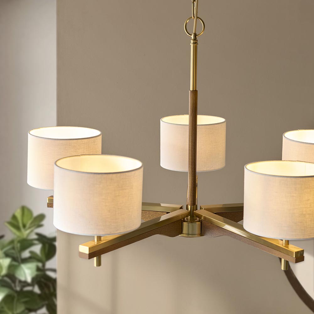 Bramwell 5 light pendant with brushed aged brass frame, oak-stained wood arms and natural linen cylinder shades, shown against a neutral wall.