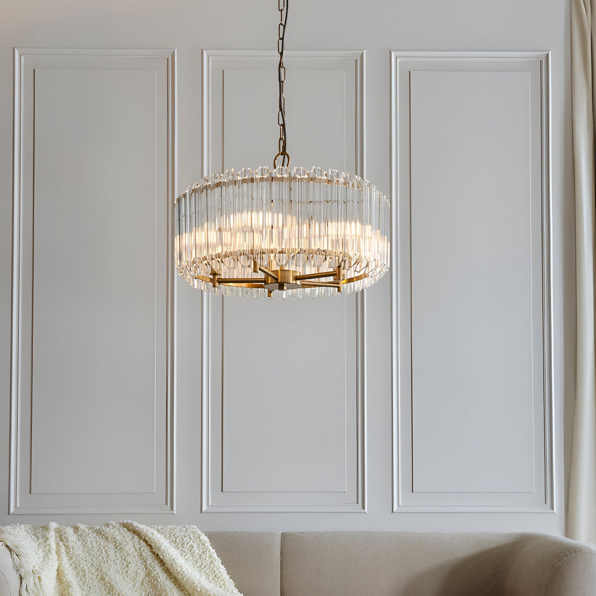 Endon Danebury 5 light pendant in brushed aged brass with clear crystal rods, hanging on a chain against a panelled wall above a cream sofa.
