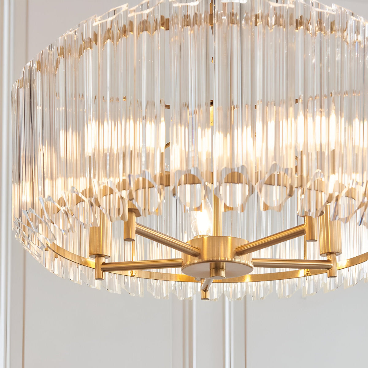 Endon Danebury 5 light pendant in brushed aged brass with vertical clear crystal rods around a drum frame, lit and shown against a pale wall.