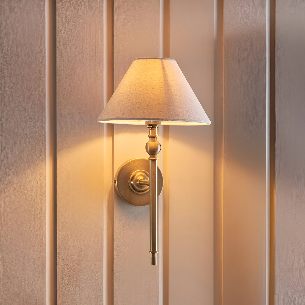 Endon Caldmere wall light in antique brass with tapered natural linen shade, mounted on a panelled wall and glowing warmly.