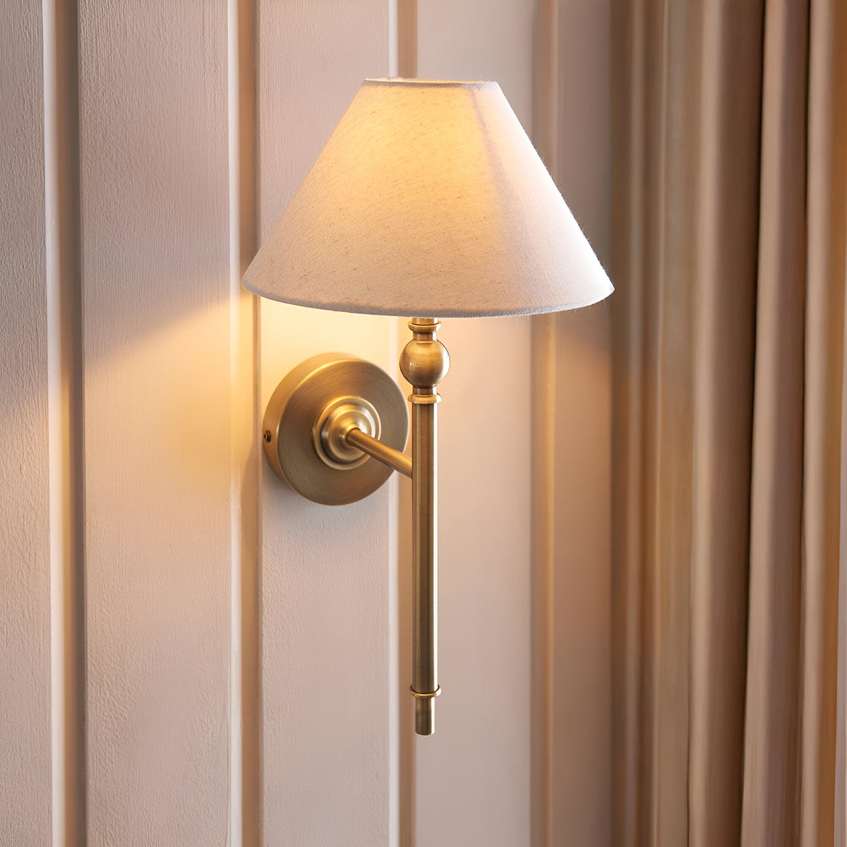 Endon Caldmere wall light in antique brass with tapered natural linen shade, mounted on a panelled wall beside soft curtains.