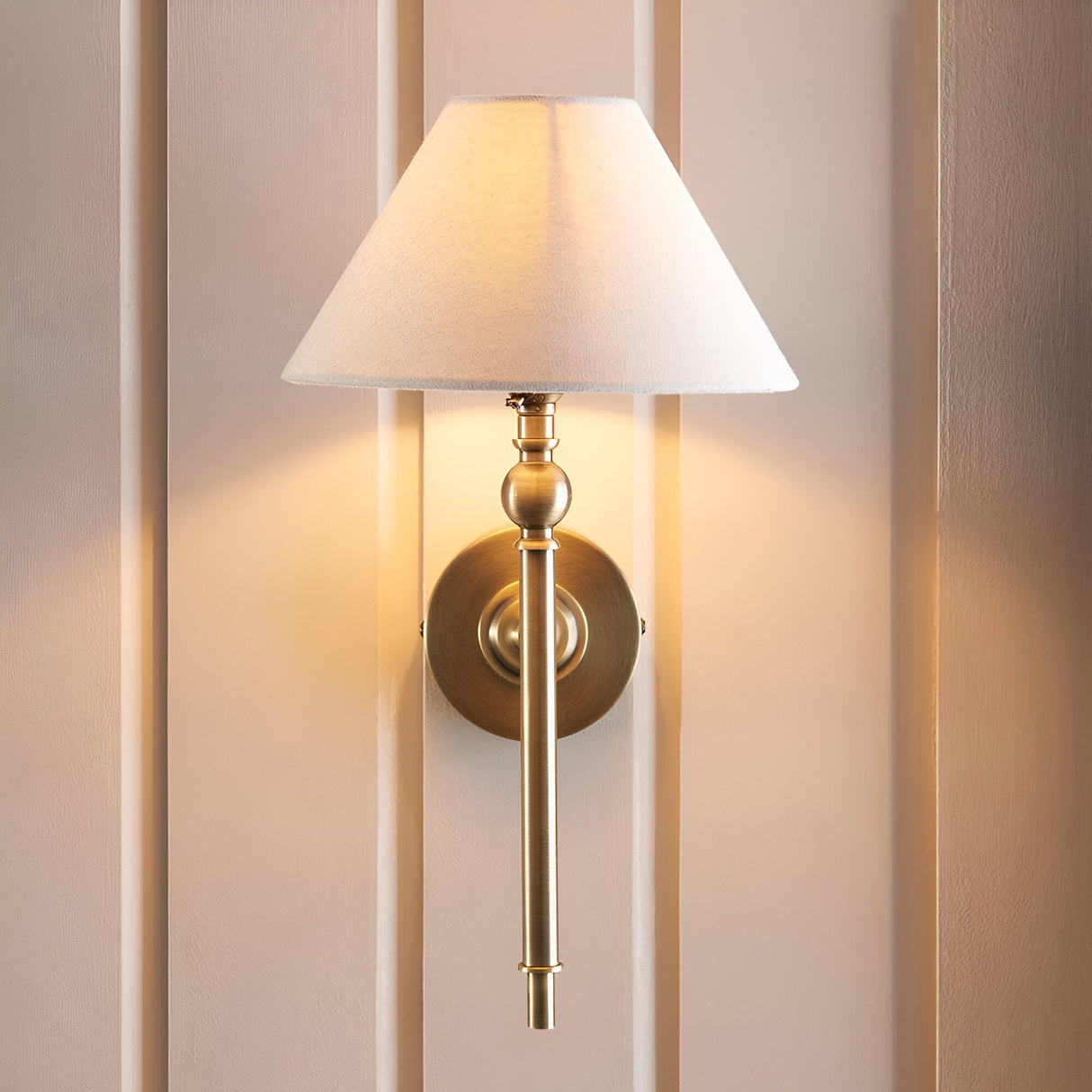 Endon Caldmere wall light in brass with tapered natural linen shade, mounted on a panelled wall casting a warm glow.