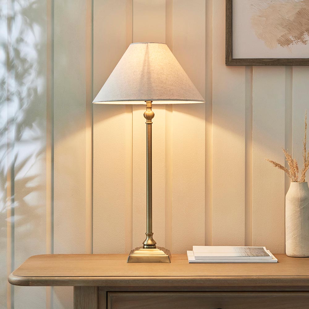 Caldmont Table Lamp in brass with a slim candlestick-style column and tapered natural linen shade, shown lit on a wooden sideboard against panelled walls.