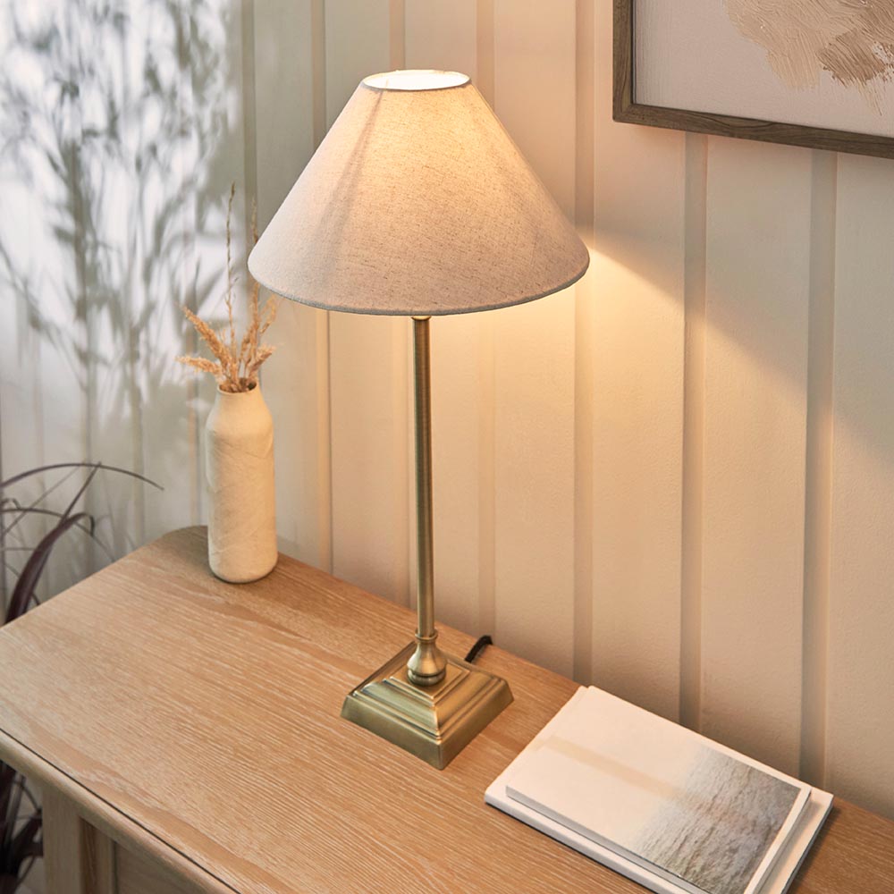 Caldmont Table Lamp in brass with a natural linen tapered shade, shown on a wooden console table beside a vase and books.