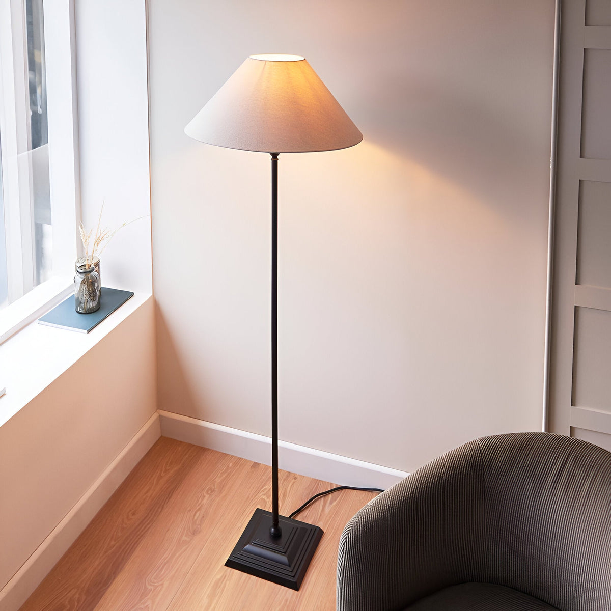 Endon Caldmere floor lamp with tapered natural linen shade and matt black metal stem on a stepped square base, beside a corduroy armchair in a bright corner.