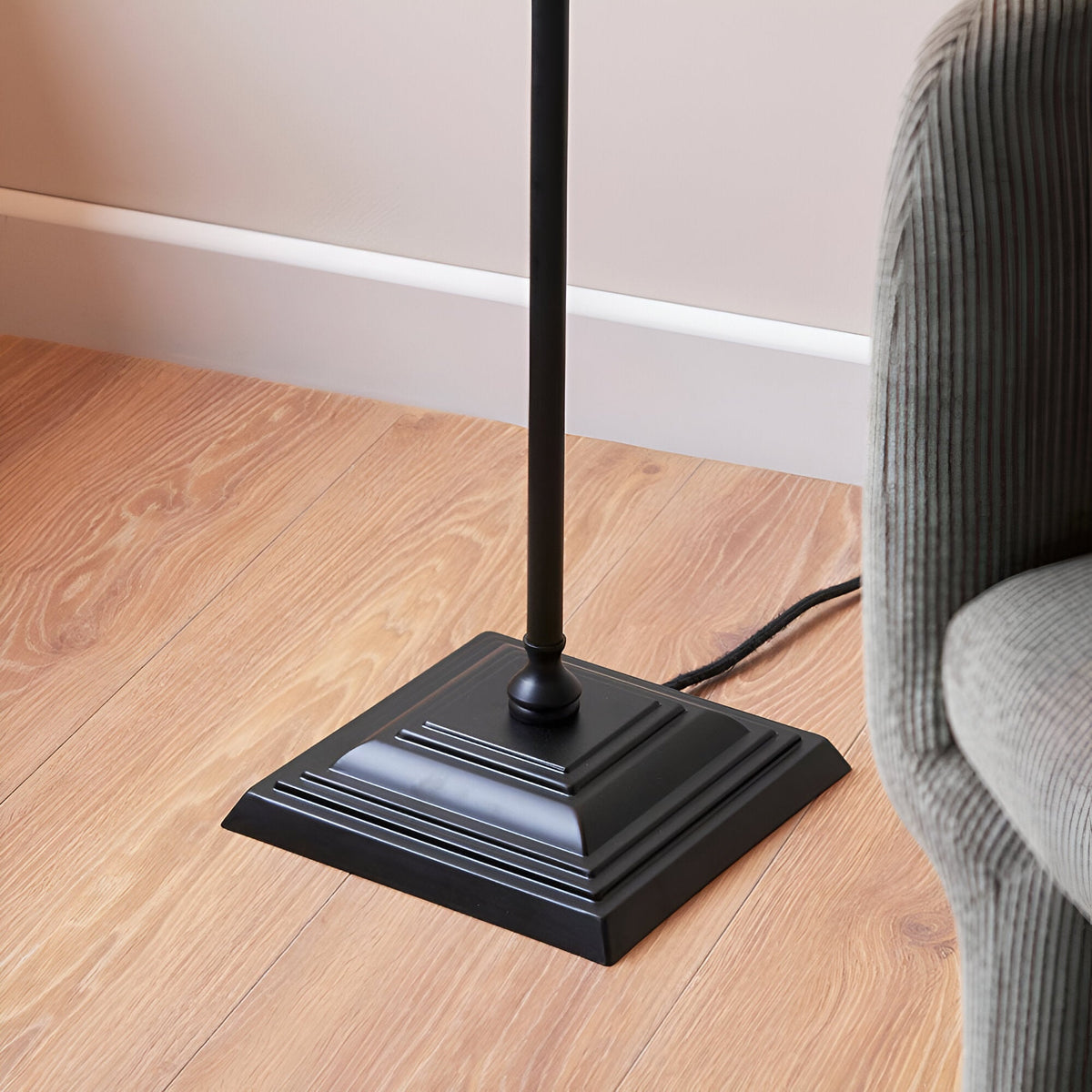 Close-up of the Endon Caldmere floor lamp's square tiered matt black metal base on wood flooring beside a grey ribbed armchair.