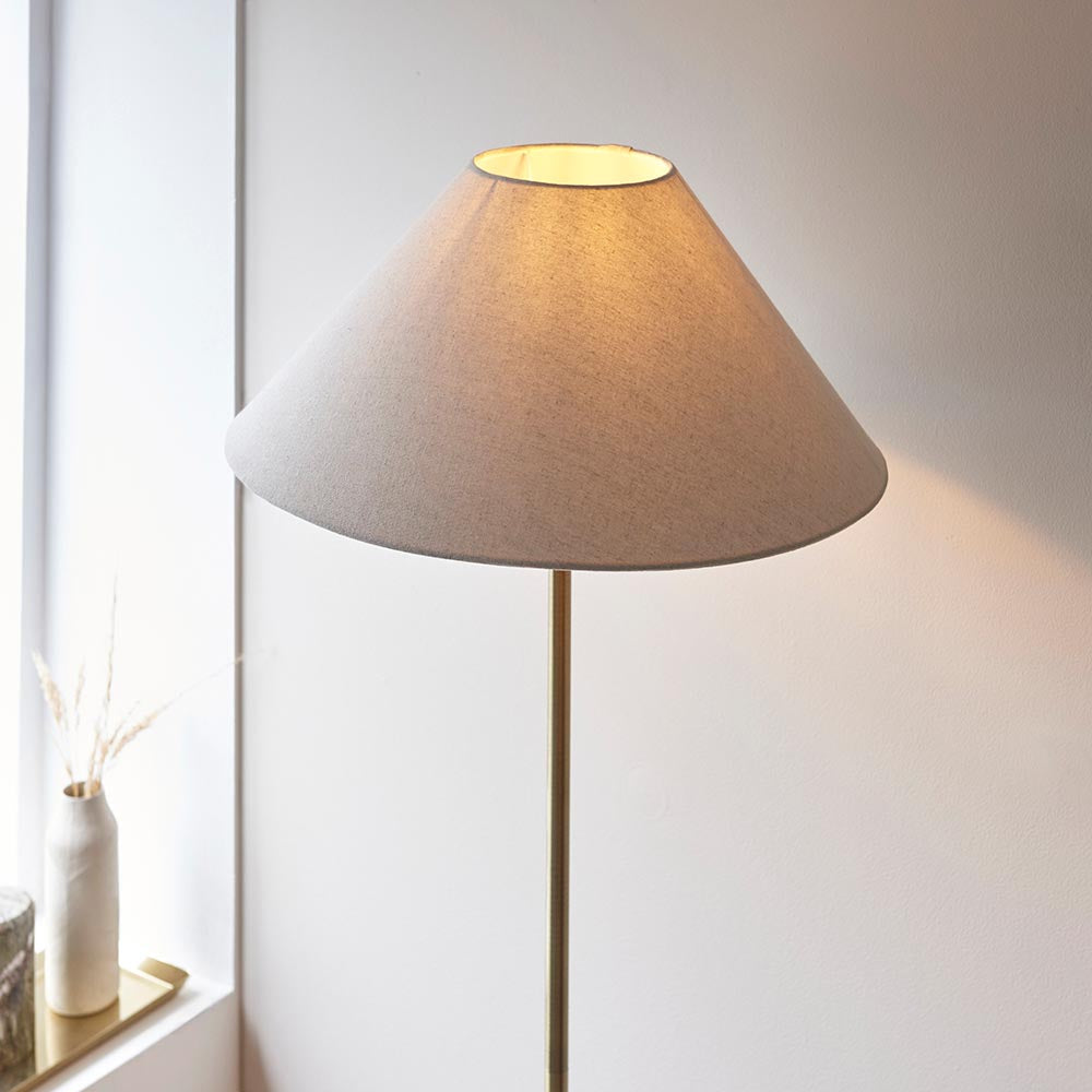 Endon Caldmere floor lamp in antique brass with a wide tapered natural linen shade, illuminated beside a windowsill with a dried grass vase.