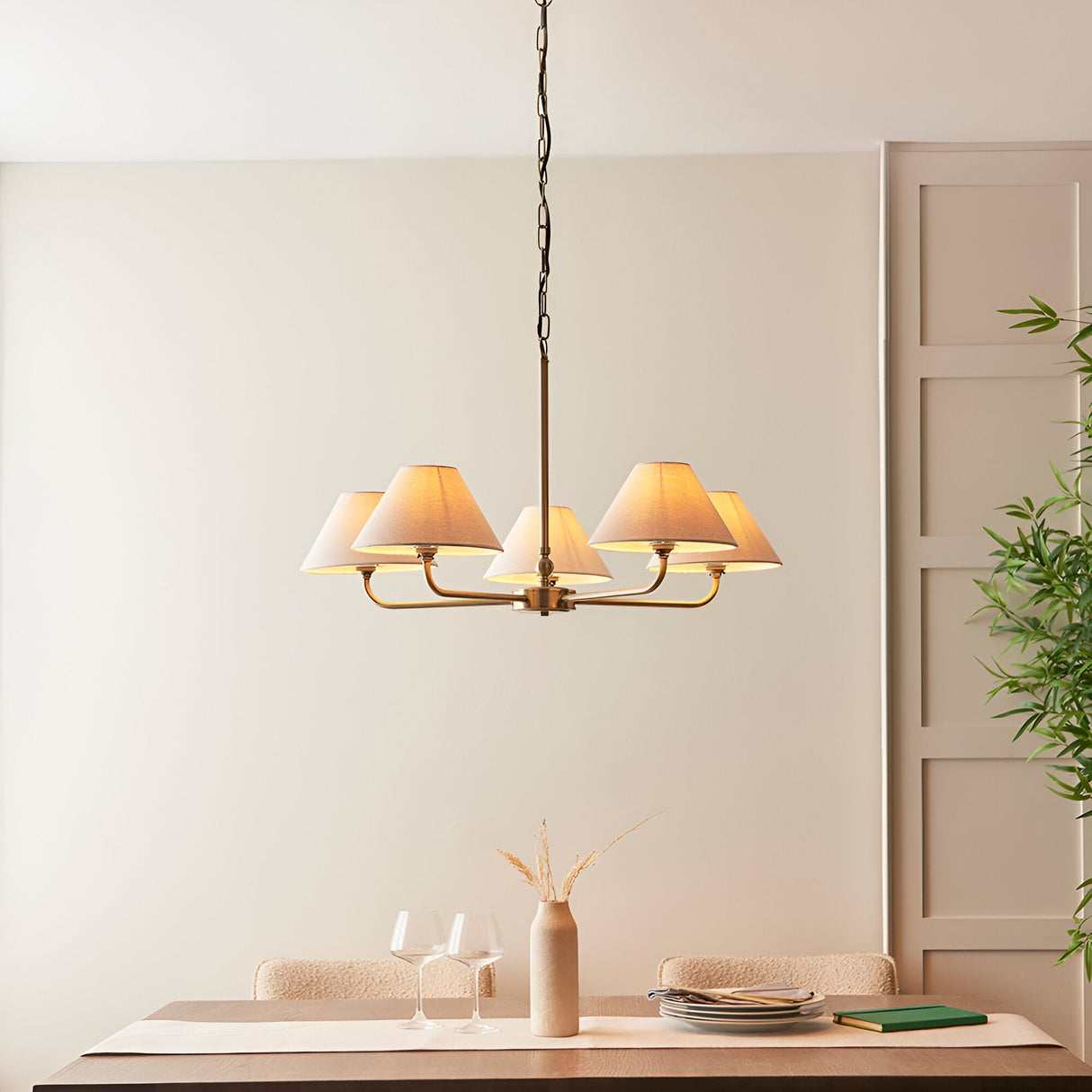 Caldmere 5 light large pendant with tapered natural linen shades on a brass finish frame and chain, shown above a dining table in a neutral room.