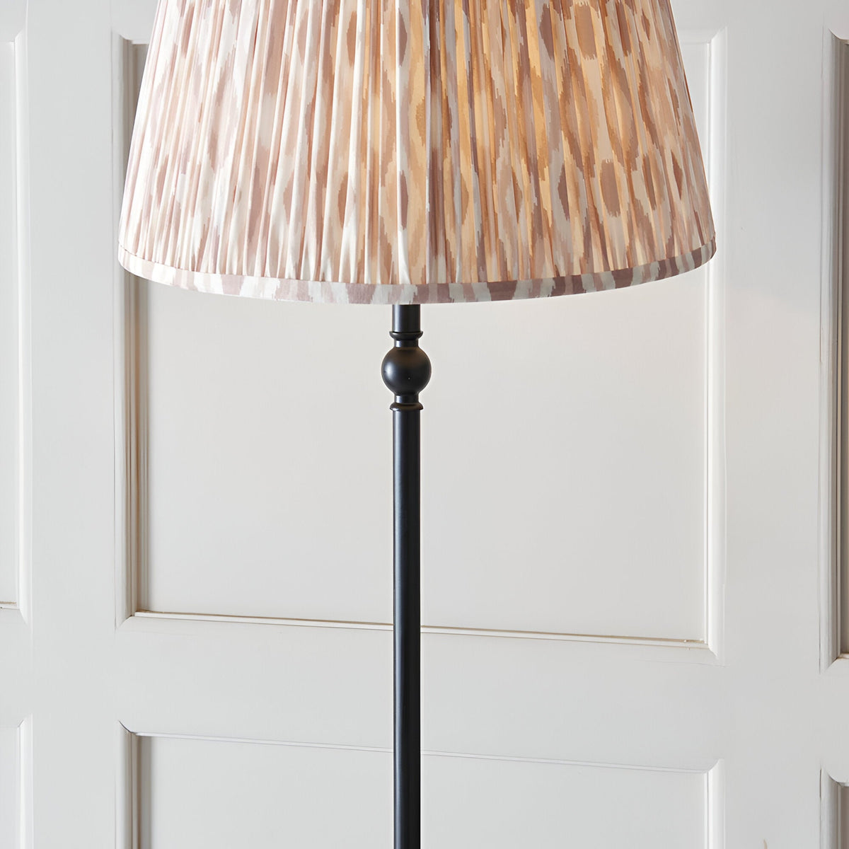 Caldmere Ikat Floor Lamp in natural fabric with a pleated 40cm shade on a matt black metal stem base, shown against a panelled white wall.