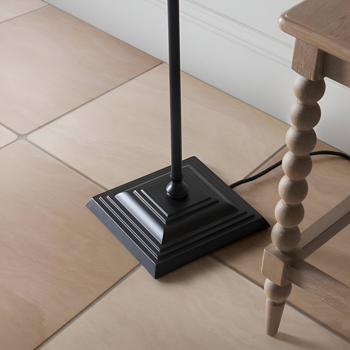 Caldmere Ikat Floor Lamp base close-up, showing matt black stepped square base and pole, on tiled floor beside a wooden table leg.