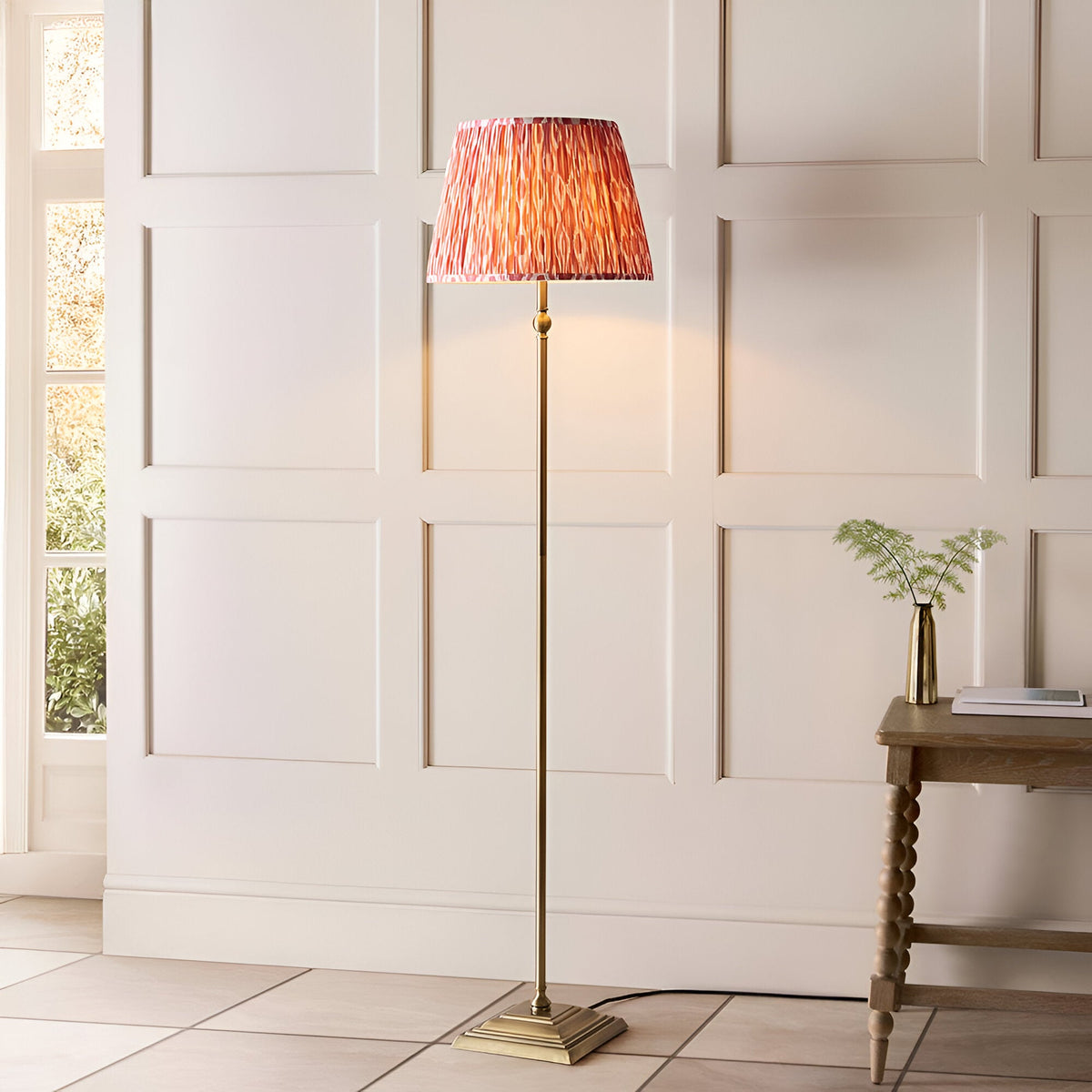 Endon Caldmere Ikat floor lamp with antique brass stand and pleated coral pink Ikat shade, placed by a panelled wall beside a wooden console table.