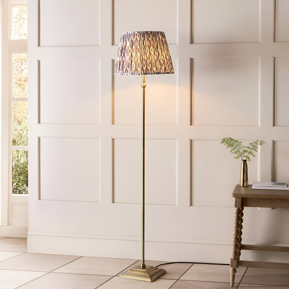 Caldmere Ikat Floor Lamp in Grey, with antique brass metal stem and square base, pleated grey patterned fabric shade, shown in a panelled living room setting.