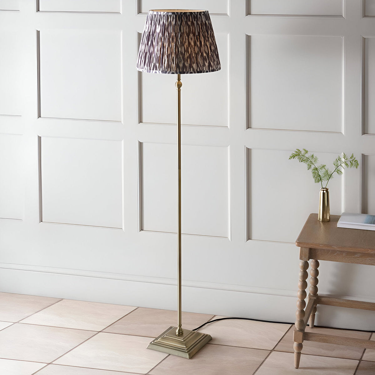 Caldmere Ikat Floor Lamp in Grey with antique brass metal stem, square base and pleated patterned shade, shown in a living room beside a wooden side table.