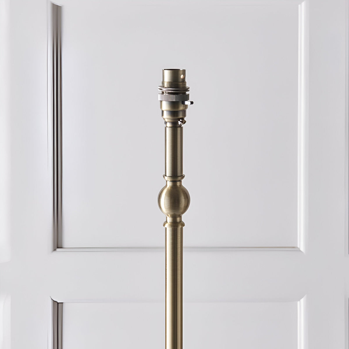 Close-up of the Endon Caldmere floor lamp's antique brass stem, showing the B22 lamp holder and turned ball detail against a white panelled door.