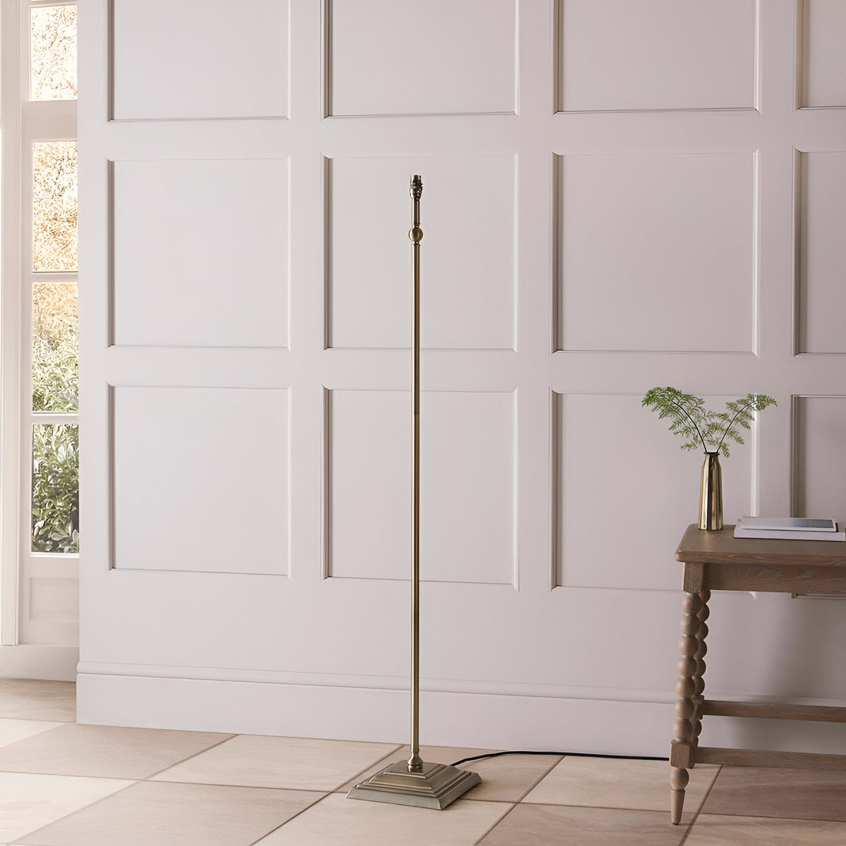 Endon Caldmere floor lamp base in antique brass with stepped square footing and B22 holder, shown without shade beside a wooden console in a panelled room.