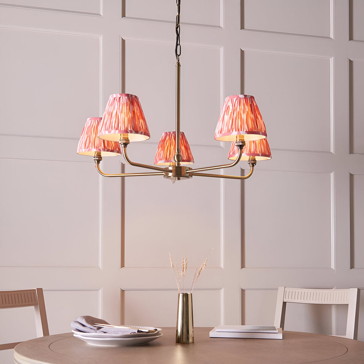 Endon Caldmere 5 light pendant with antique brass arms and coral pink pleated ikat tapered shades, hung above a dining table against panelled walls.