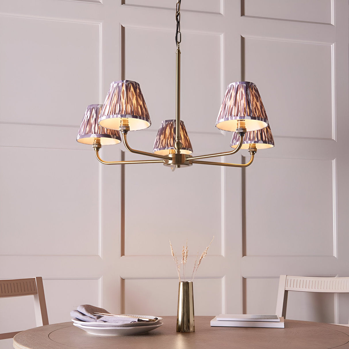 Caldmere 5 Light Ikat Pendant in antique brass with grey pleated fabric tapered shades, hanging above a dining table in a light-panelled room.