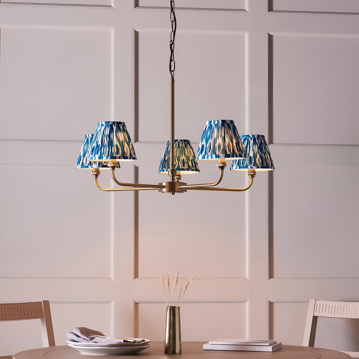 Endon Caldmere 5 light pendant with tapered pleated marlin blue ikat fabric shades on antique brass arms, hung above a dining table.