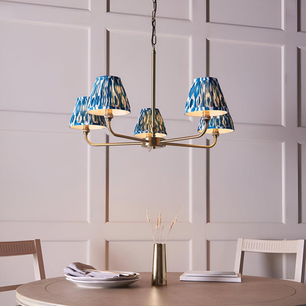 Endon Caldmere 5 light pendant in antique brass with tapered pleated marlin blue ikat fabric shades, hung above a dining table against panelled walls.