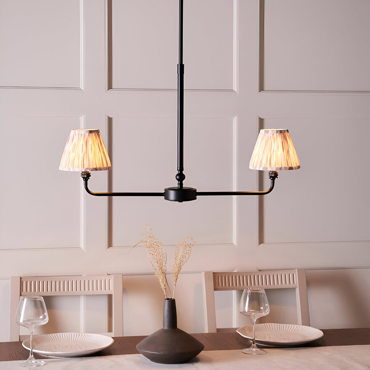 Endon Caldmere 2 light linear pendant in matt black with tapered natural pleated fabric shades, shown above a dining table against a panelled wall.