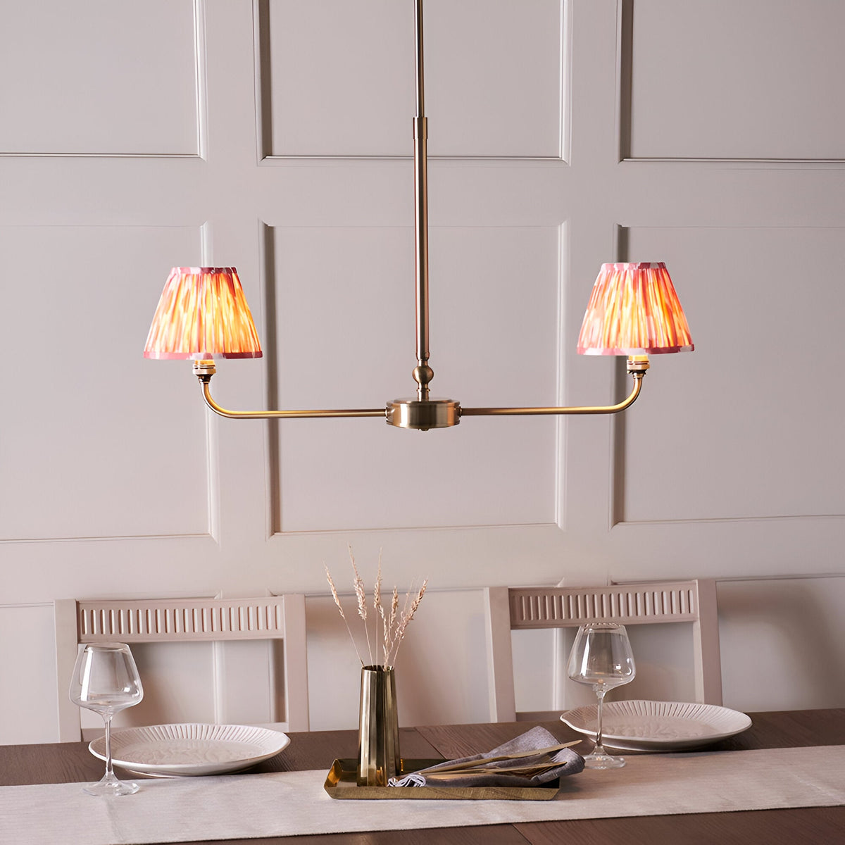Endon Caldmere 2 light linear pendant in antique brass with coral pink pleated ikat tapered shades, shown above a dining table set with glassware.
