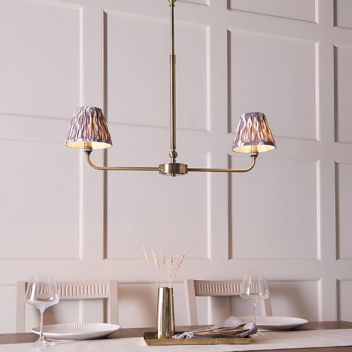 Caldmere 2 Light Ikat Pendant in antique brass with grey pleated tapered shades, shown above a dining table in a panelled room.