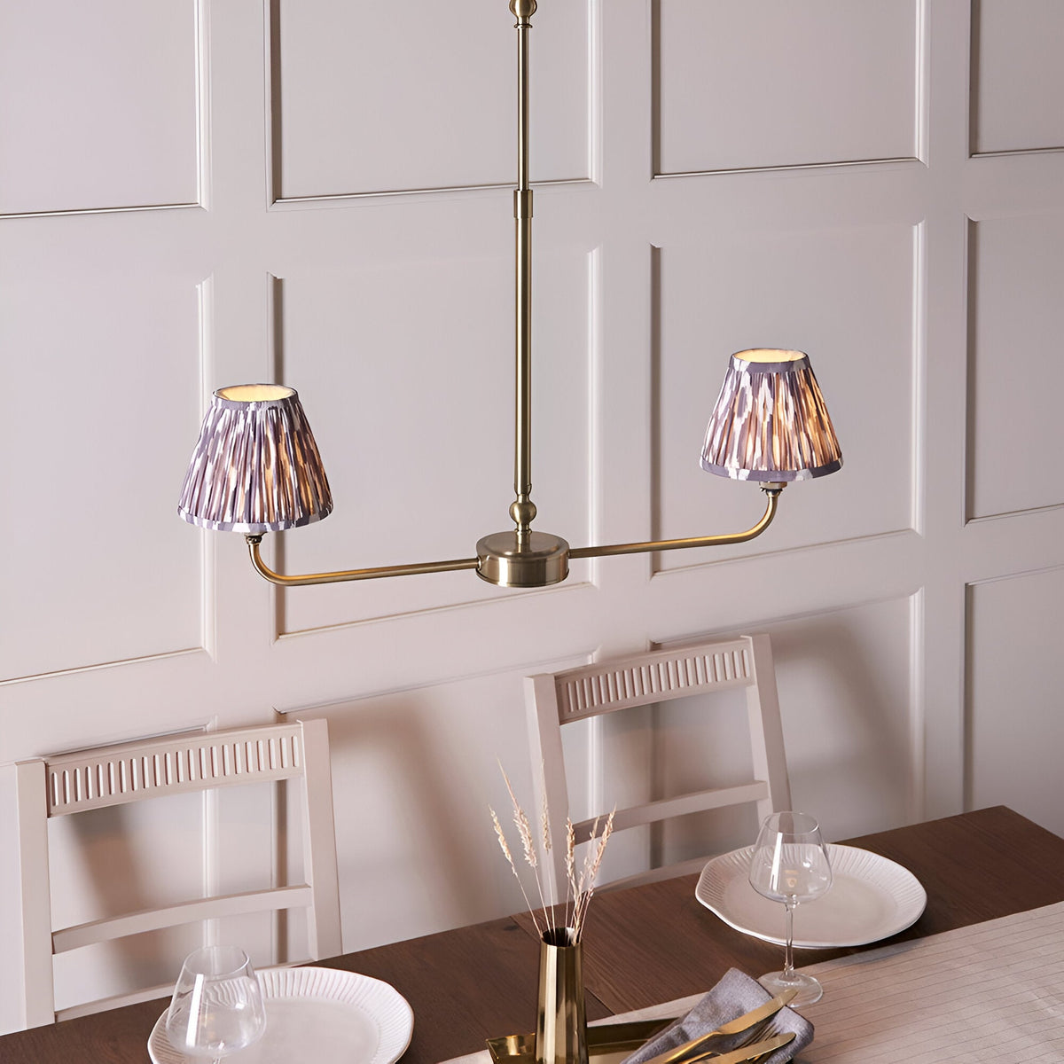 Caldmere 2 Light Ikat Pendant in antique brass with grey pleated tapered shades, shown above a dining table against panelled walls.