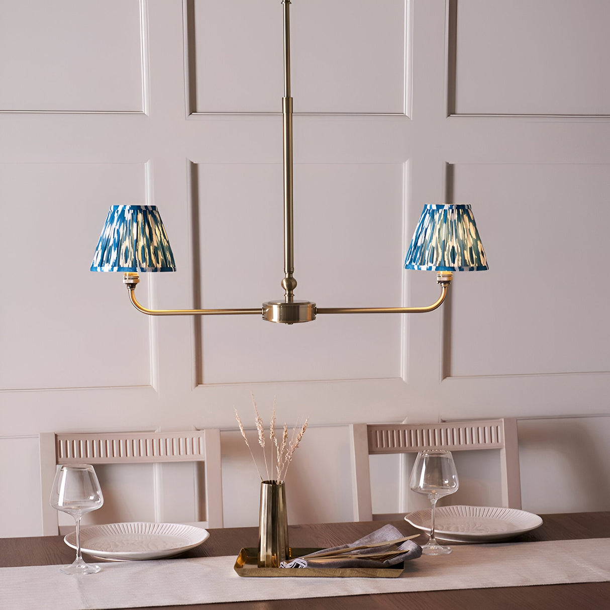 Endon Caldmere 2 light pendant in antique brass with twin tapered blue ikat pleated shades on a linear frame, shown above a dining table.