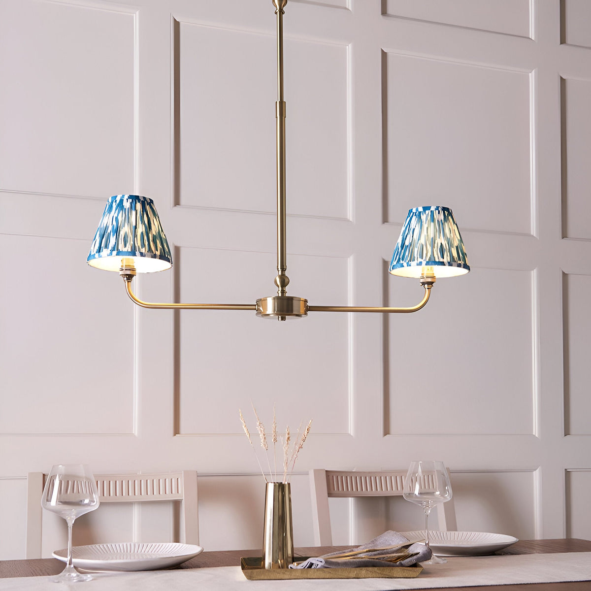 Endon Caldmere 2 light pendant with tapered blue ikat fabric shades on an antique brass linear frame, shown above a dining table.