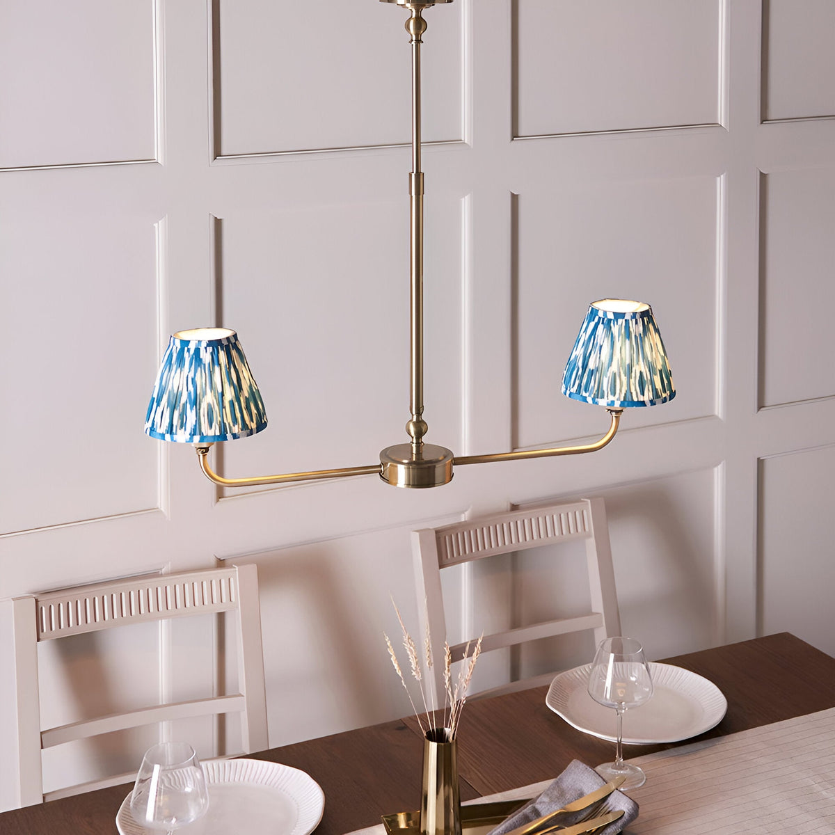 Endon Caldmere 2 light pendant in antique brass with tapered blue ikat pleated shades, hanging above a dining table against a panelled wall.