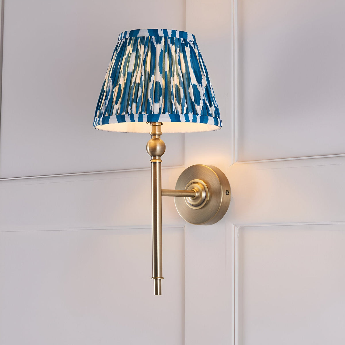 Caldmere Ikat Wall Light in Blue with pleated tapered fabric shade on an antique brass arm and round wall mount, shown against a panelled white wall.