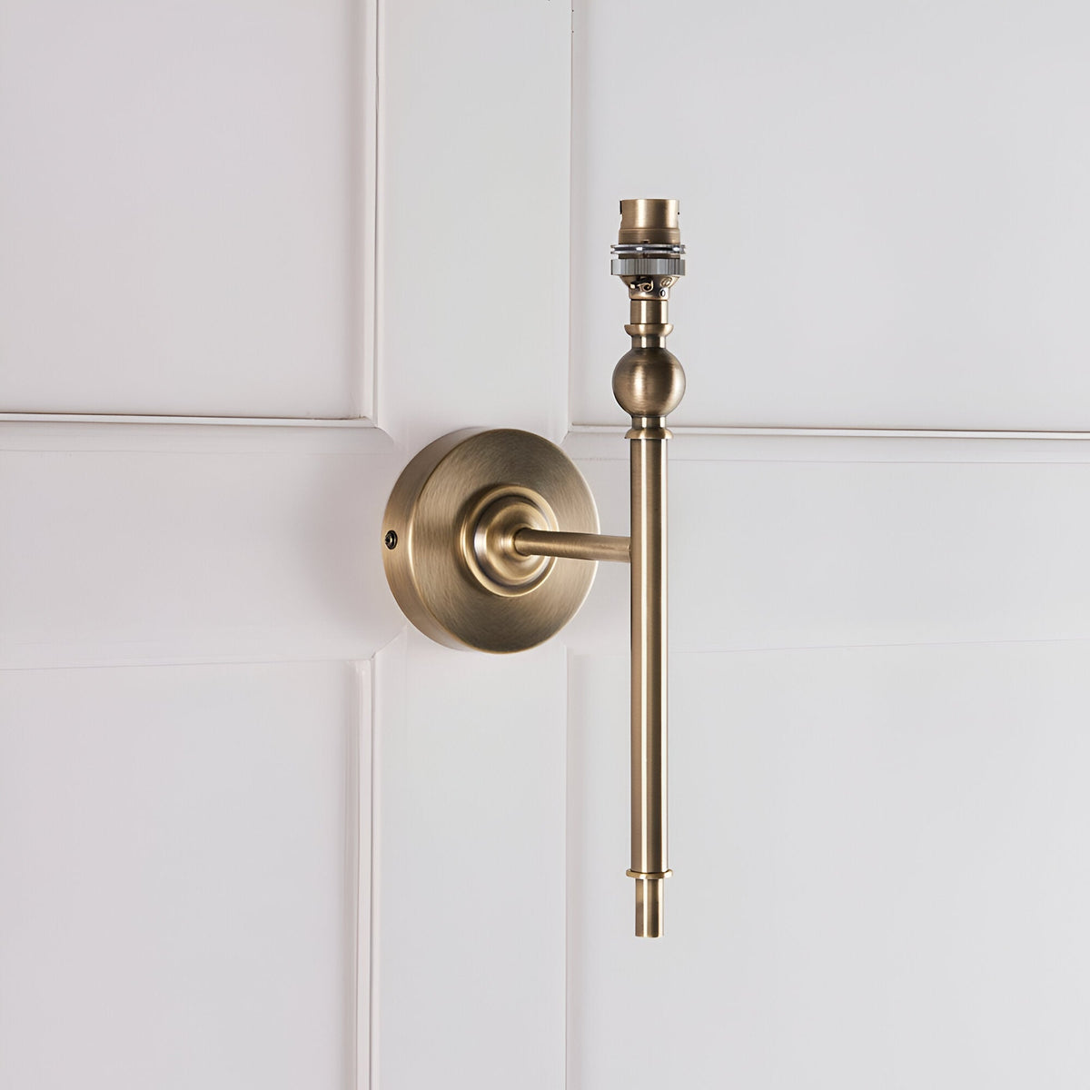 Caldmere antique brass wall light with round backplate, straight arm, and B22 lamp holder, shown without shade against a white panelled wall.