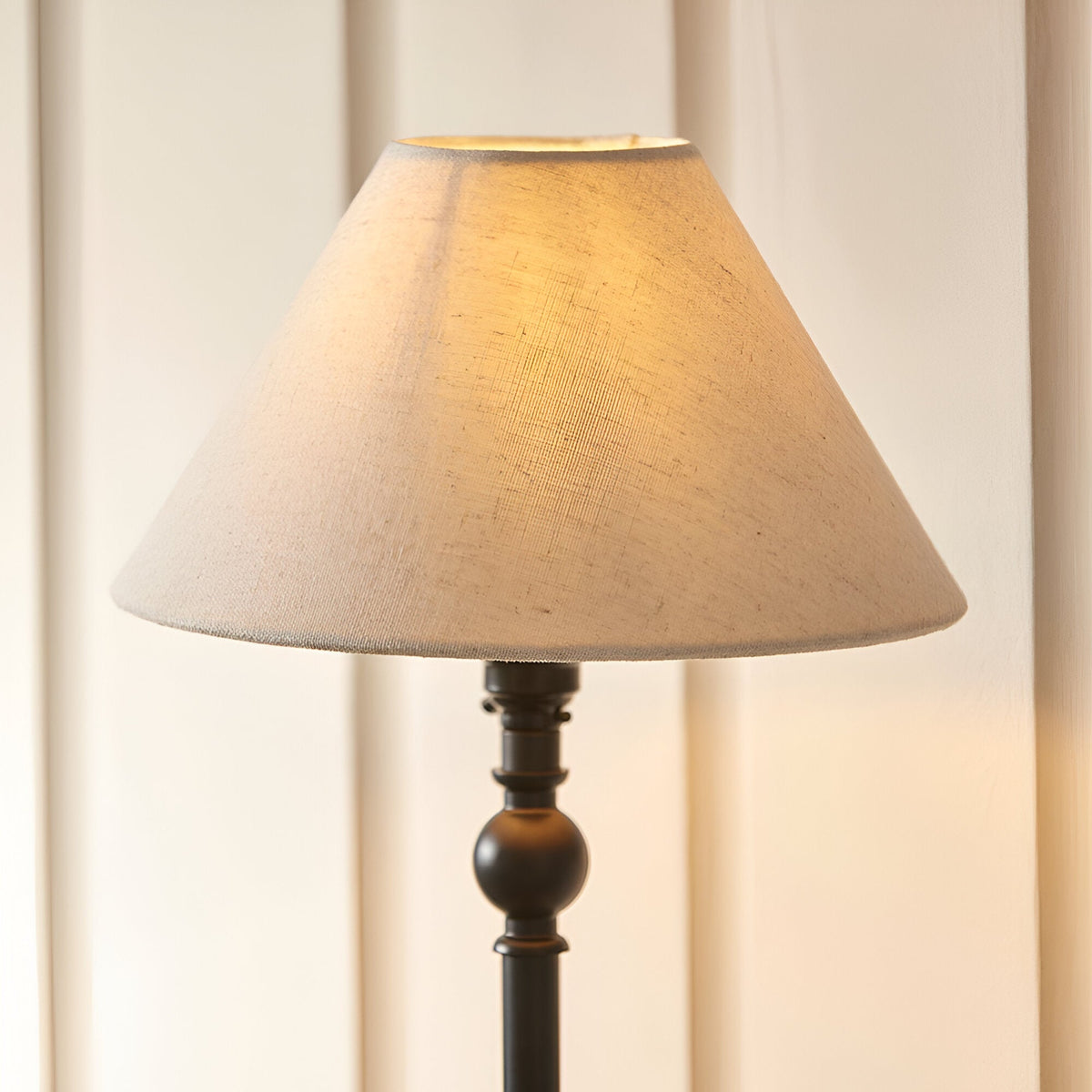 Endon Caldmont table lamp with matt black candlestick base and tapered natural linen mix shade, illuminated against a pale panelled wall.