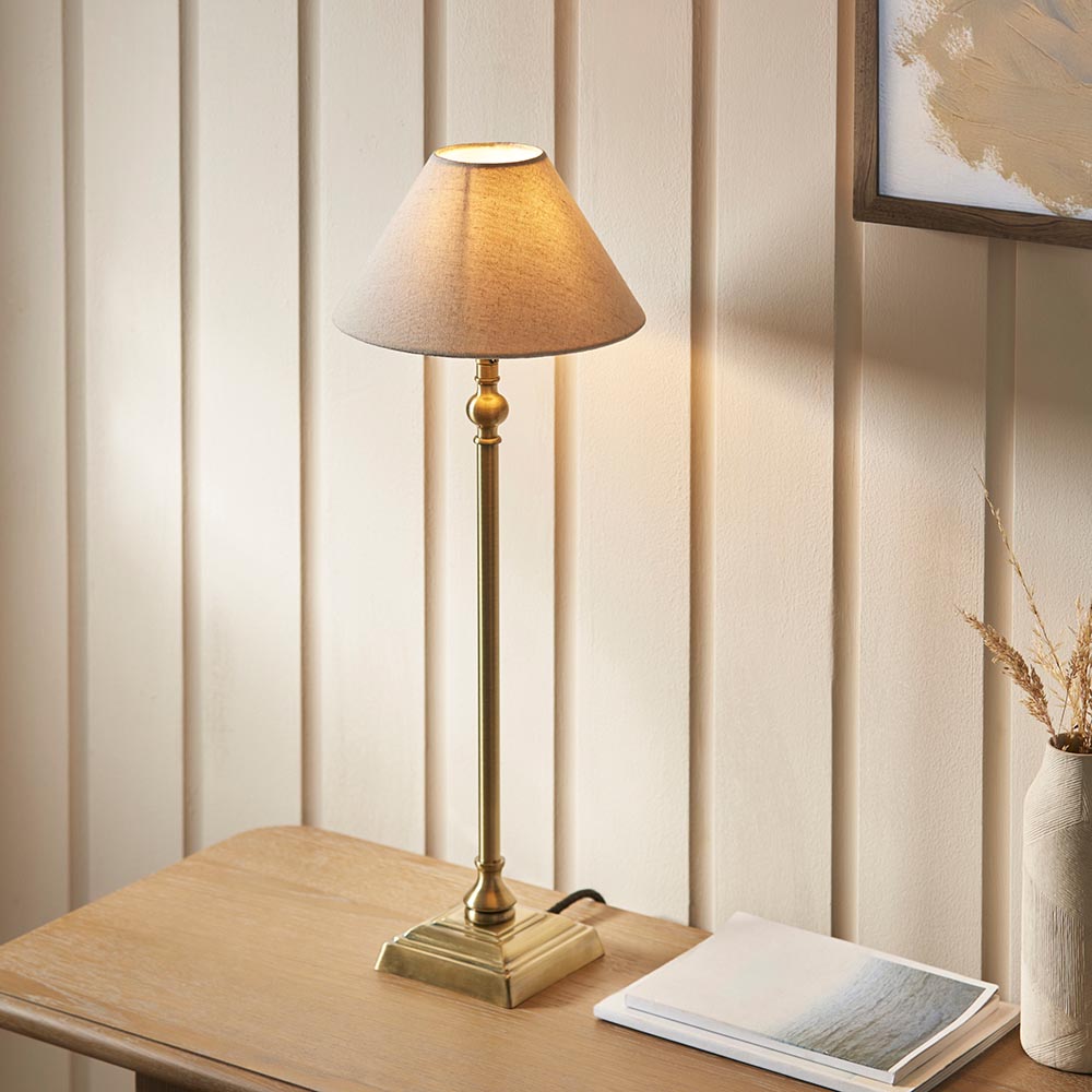 Endon Caldmont table lamp in antique brass with a slim candlestick column and tapered natural linen shade, lit on a wooden side table by a panelled wall.