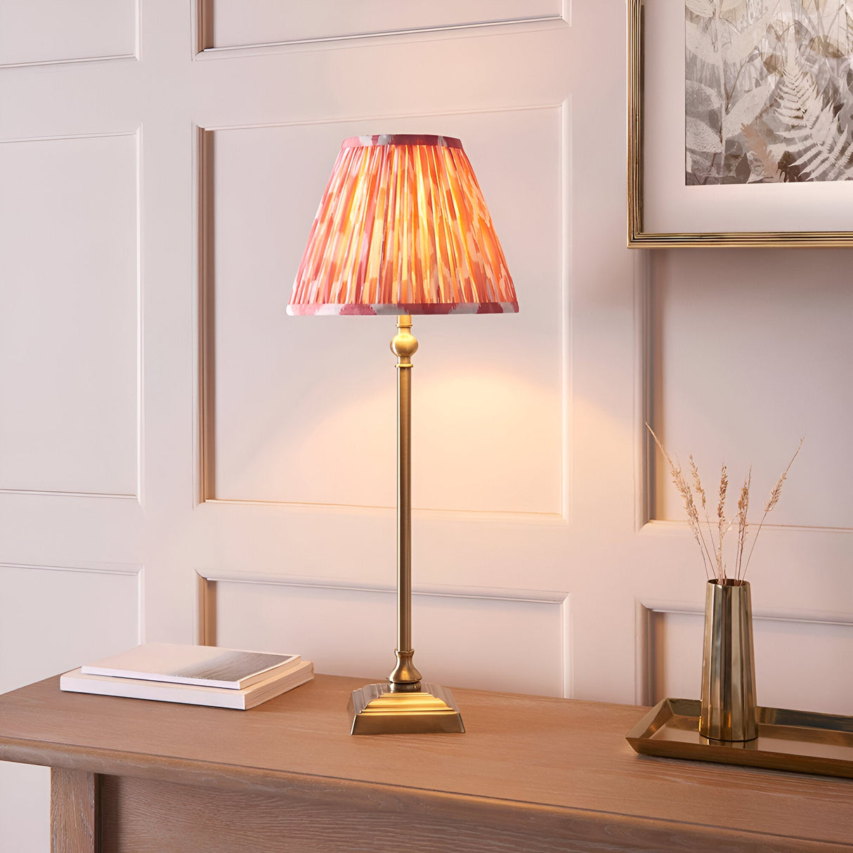 Caldmere Ikat Table Lamp in Coral, an antique brass candlestick-style base with a pleated coral fabric shade, shown lit on a wooden console table.
