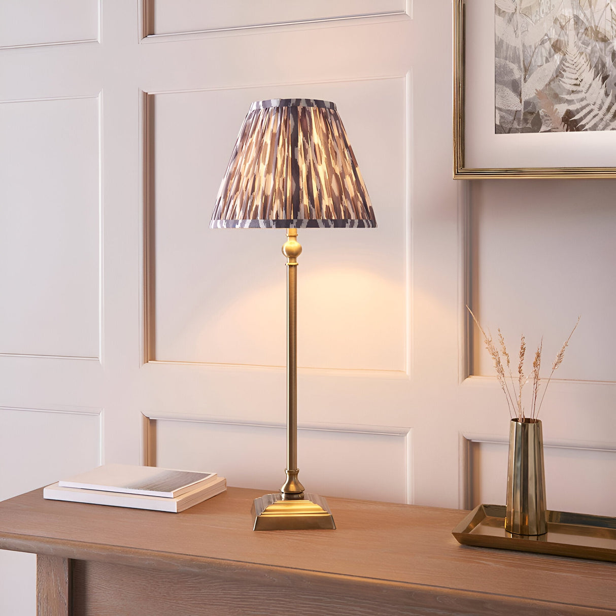 Endon Caldmere table lamp with slim antique brass candlestick stem and pleated grey Ikat print shade, styled on a wood console beside books and a brass vase.