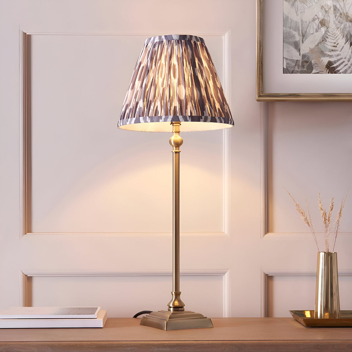 Endon Caldmere table lamp with slim antique brass candlestick stem and pleated grey Ikat print shade on a wooden sideboard against panelled walls.
