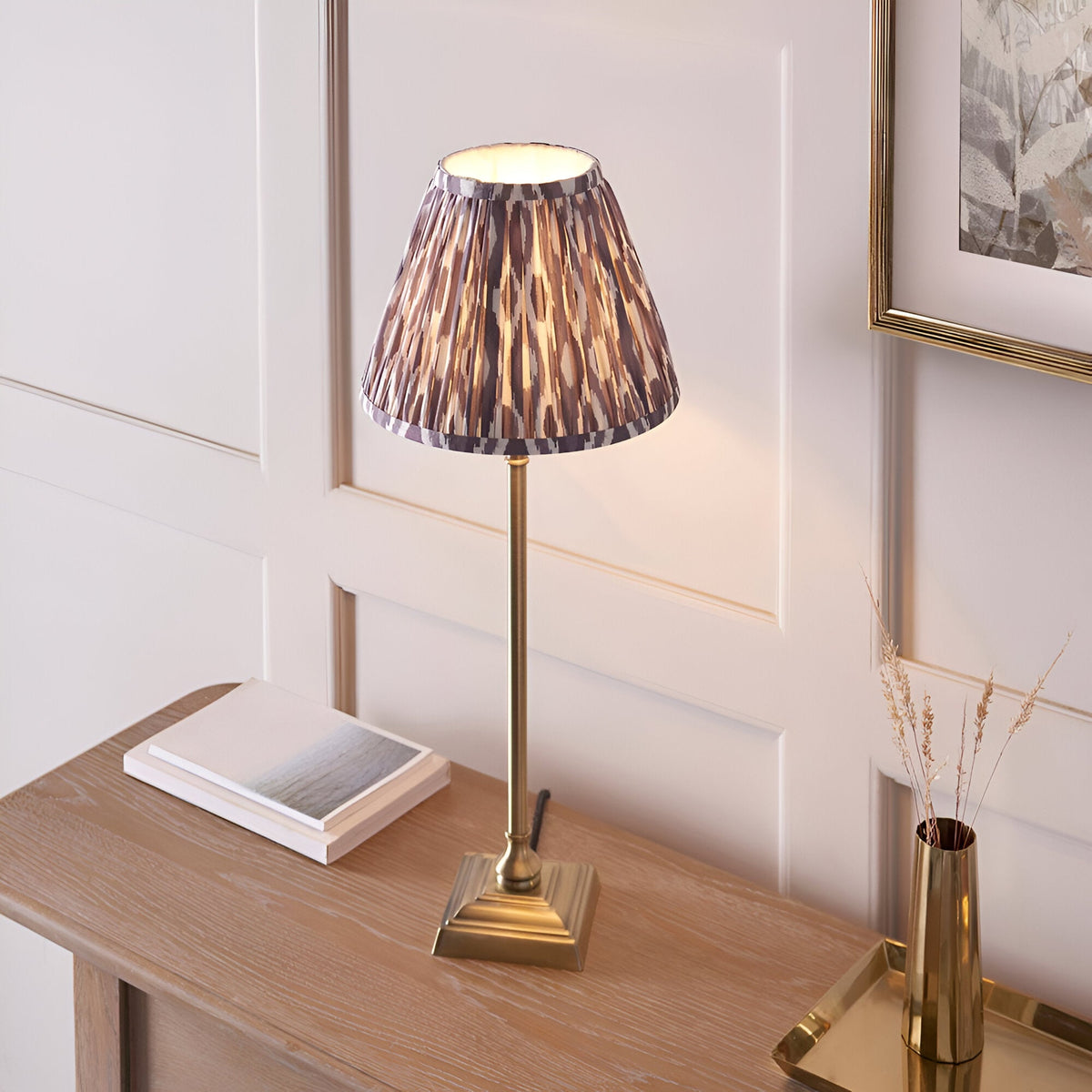 Endon Caldmere table lamp with slim antique brass candlestick base and pleated grey Ikat print shade, on a wood console beside books and a brass vase.