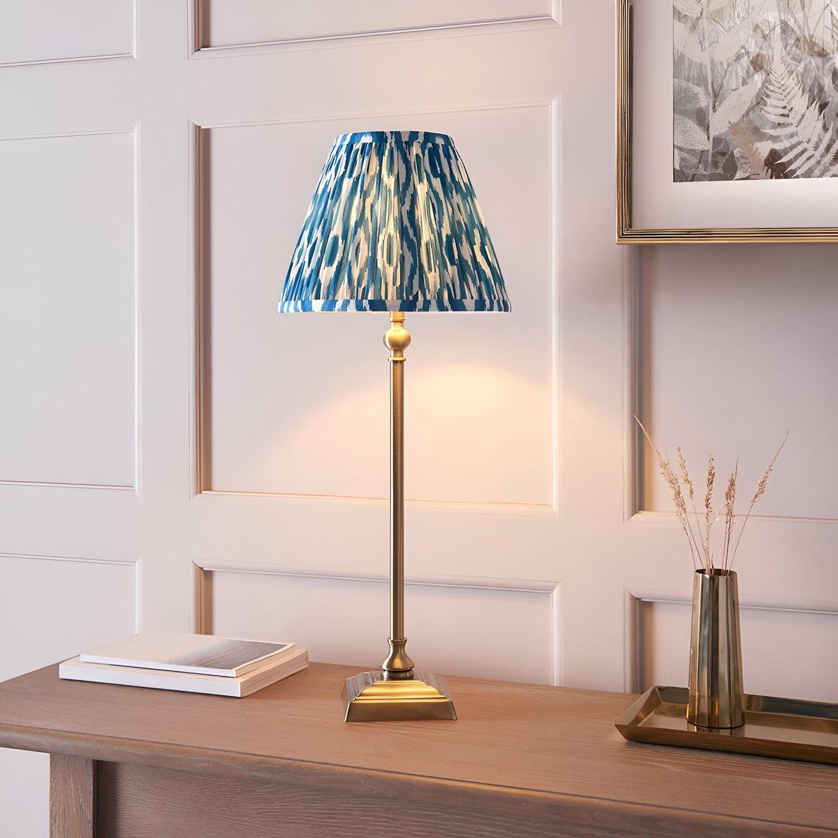 Endon Caldmere table lamp with slim antique brass candlestick stand and pleated blue Ikat shade, on a wood console beside books and a brass vase.
