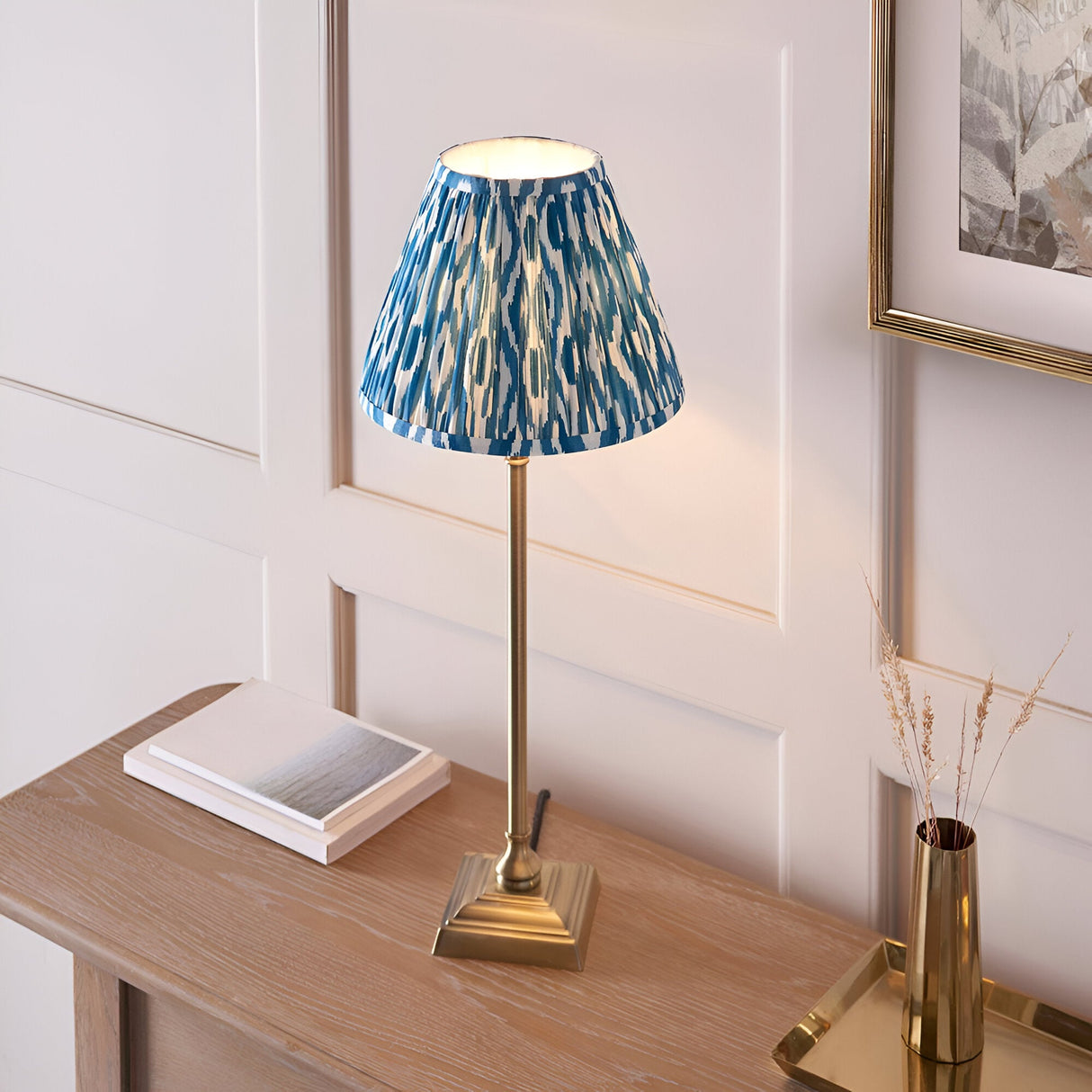Endon Caldmere table lamp with slim antique brass candlestick base and pleated blue Ikat shade, on a wood console beside books and a brass vase.