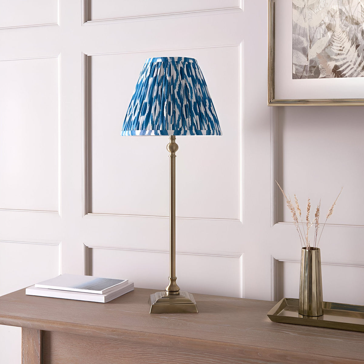 Endon Caldmere table lamp with slim antique brass candlestick base and pleated blue Ikat shade, on a wood console beside books and a brass vase.
