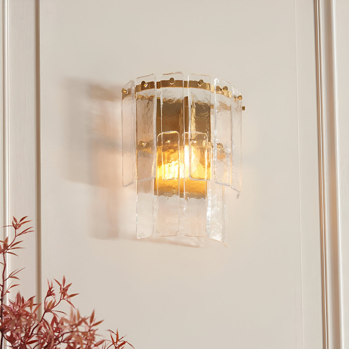 Brockhurst Wall Light in Brass, featuring layered textured glass panels on a brushed aged brass backplate, illuminated on a light wall beside foliage.