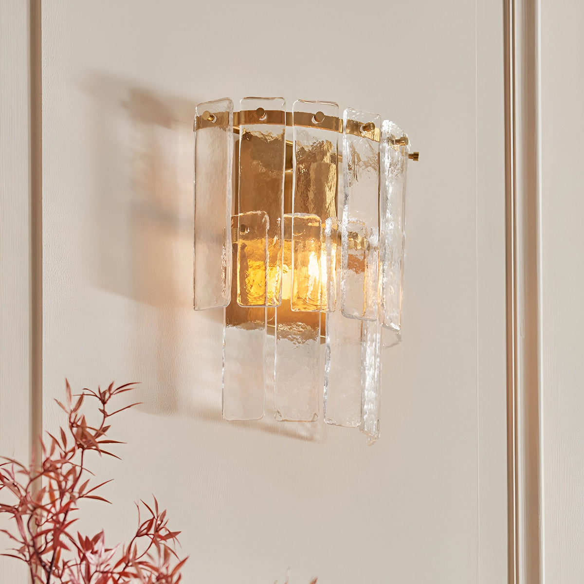 Brockhurst Wall Light in Brass, layered textured glass panels on a brass backplate, illuminated on a light wall beside red foliage.