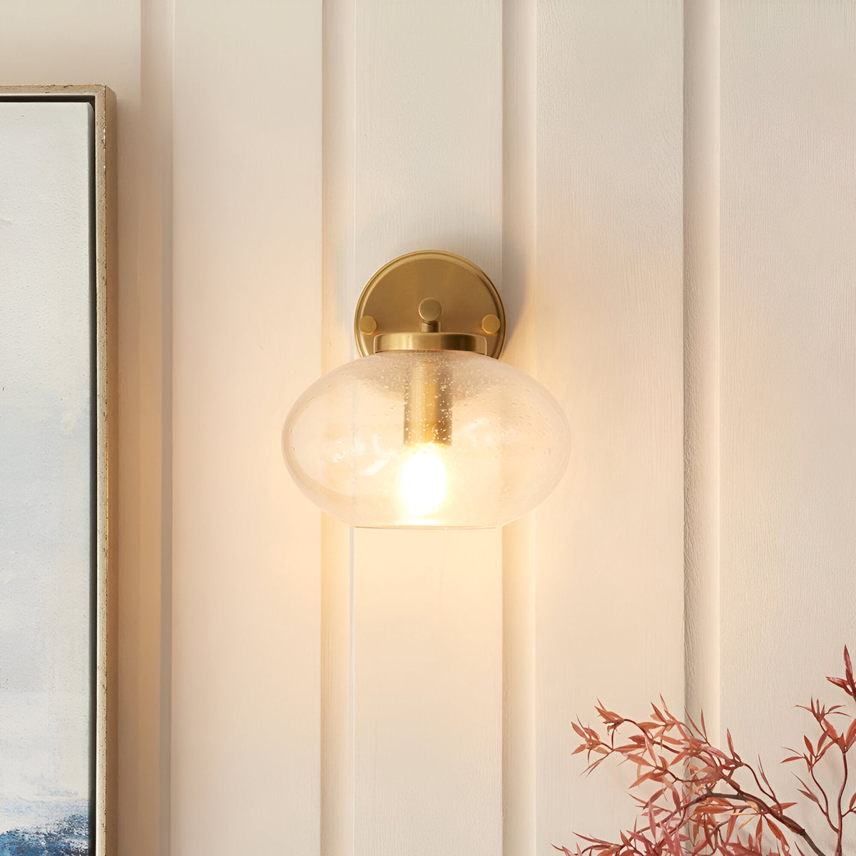 Endon Pemberton wall light in brass with a bubbled clear glass oval shade, illuminated on a panelled cream wall beside framed art and foliage.
