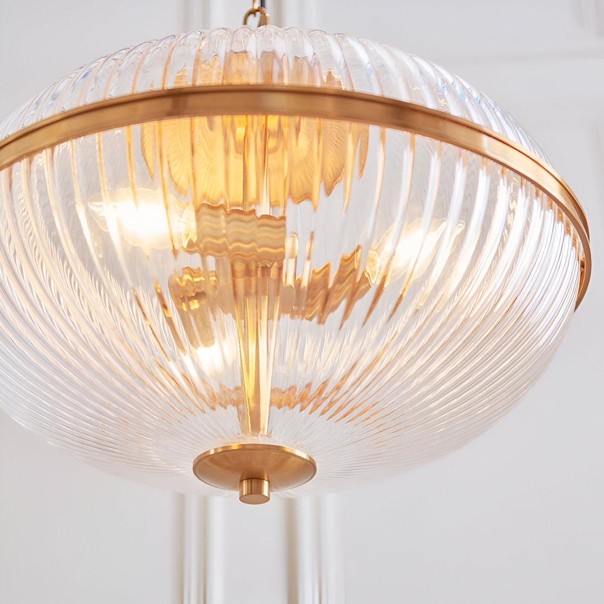 Langton 3 Light Pendant in Brass, close-up of the ribbed two-part clear glass globe held by a shaped brass band, lit from within against a white interior.